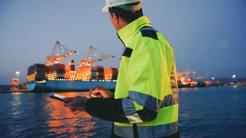 Foreperson in protective gear using digital tablet in front of container terminal port during night